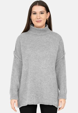 Woman with long brown hair wearing gold hoop earrings and a loose gray turtleneck sweater, standing against a plain light background.