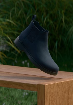 Bota corta azul oscuro sin cordones flotando sobre un banco de madera con césped verde y plantas al fondo.