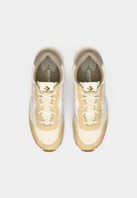 Beige sneakers med struktureret stof, hvide læderdetaljer og en brun gummisål. Har rundt logo på indersålen og gulddetaljer på tungen.