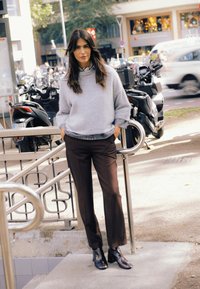 Pulover supradimensionat gri deschis cu mâneci lungi; pantaloni maro, croiți; ghete negre până la gleznă; decor urban cu motociclete în fundal.