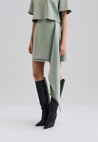 Mintgrünes Kleid mit lockerer Passform, asymmetrischem Saum und drapiertem Akzent. Kombiniert mit schwarzen knielangen Stiefeln mit spitzer Zehenpartie.