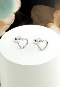 Silverhjärtformade örhängen med pärlmönster, med en genomskinlig baksida för säker användning. Uppställda på en vit, cirkulär displayställ.