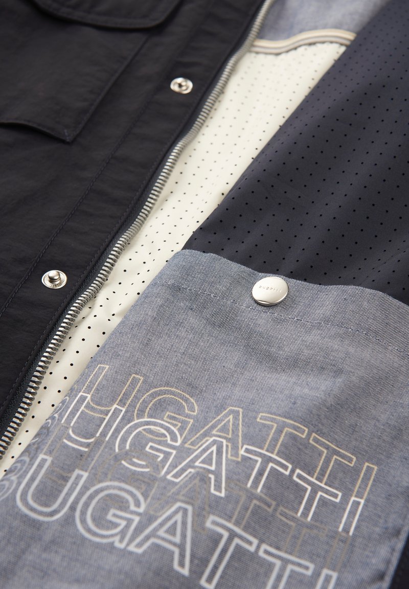 Herrenjacke mit navyfarbener Außenseite, Reißverschluss, kontrastierendem Innenfutter mit cremefarbenen und navyblauen Punkten sowie einem strukturierten blauen Paneel mit einem Logo.