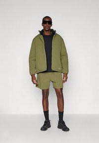 Olive green padded jacket with zipper and high collar, paired with matching shorts. Black t-shirt underneath, black sunglasses, and sporty shoes.