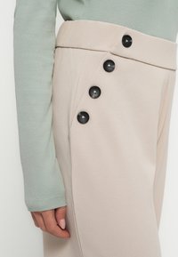 Beige tailored broek met een hoge taille, met een gladde textuur en zes zwarte knopen langs de zijkant. Een mouw van een muntgroene geribbelde trui is zichtbaar.