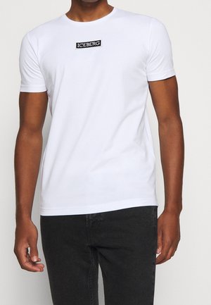 Mężczyzna ubrany w gładki biały T-shirt z krótkim rękawem z czarno-białą naszywką z logo "ICEBERG" na klatce piersiowej oraz czarne spodnie.