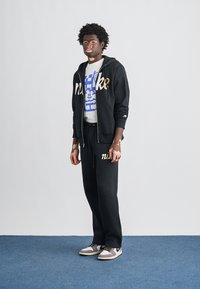 Fekete kapucnis pulóver és melegítőnadrág, mindkettőn arany "nike" logóval. Alatta fehér mintás póló kék dizájnnal. Szürke és fehér sportcipők.