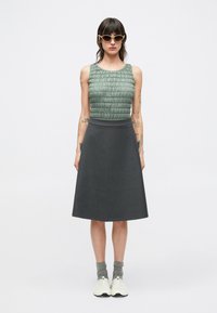 Groen, gestructureerd mouwloos topje gecombineerd met een donkergrijze uitlopende rok. Het model draagt witte sneakers en grijze sokken, met een zonnebril.