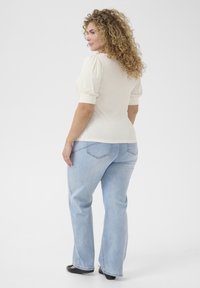 Haut crème à manches bouffantes, associé à un jean droit bleu clair. Le mannequin se tient légèrement de côté, avec des cheveux bouclés.