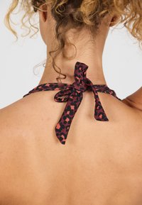 Halter neck in tessuto nero con un motivo leopardato in rosso e rosa, fissato con un fiocco annodato sul retro, che rivela una pelle liscia.