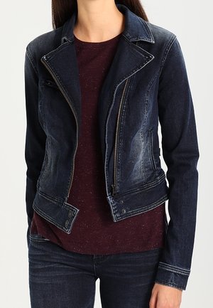Femme portant une veste en jean foncé ajustée sur une chemise bordeaux mouchetée avec un jean en denim foncé assorti.