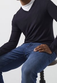 Granatowy sweter w serek z długimi rękawami, zestawiony z niebieskimi dżinsami. Zbliżenie na siedzącą modelkę, ukazujące fakturę materiału i ułożenie rąk.
