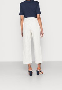 Pantalon large blanc à fines rayures, taille haute avec deux poches arrière, associé à un T-shirt bleu marine et des sandales à talons avec des accents orange.