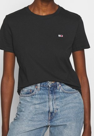Person wearing a black short-sleeve t-shirt with a small, rectangular red, white, and blue logo on chest and light blue jeans.