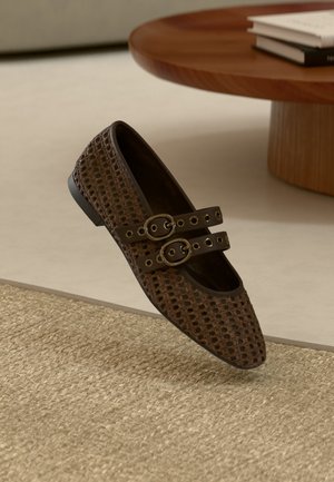 Dunkelbraunes geflochtenes Leder-Ballettschuh mit zwei Riemen und Schnallen, platziert neben einem runden Holztisch und einem beigen gewebten Teppich.