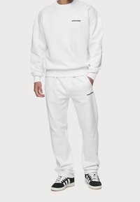 Bílá mikina a odpovídající kalhoty, obojí vyrobené z měkké tkaniny, mají černé logo "PEGADOR". Kalhoty mají elastický pas.