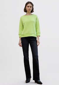 Sudadera de color verde claro con mangas largas, cuello amplio y corte relajado, combinada con jeans negros acampanados y botines.