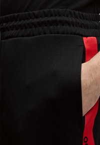 Χέρι στην τσέπη μαύρου παντελονιού με ελαστική μέση και κόκκινη κάθετη ρίγα στο πλάι.