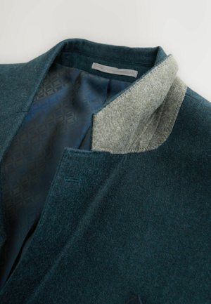 Grüne Wolljacke mit einem weichen grauen Kragen. Das Innenfutter hat ein gemustertes dunkelblaues Futter. Enthält eine gestickte Detail am Kragen.