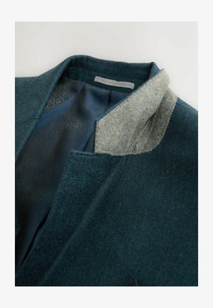 Grøn uldjakke med en blød grå krave. Indersiden har et mønstret mørkeblåt for. Inkluderer en syet detalje ved kraven.