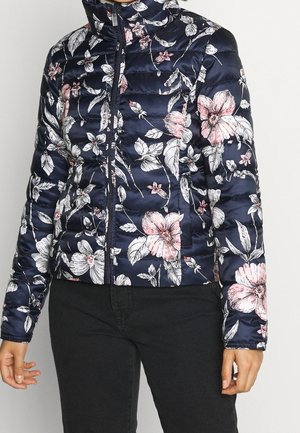 Chaqueta acolchada azul marino con estampado floral rosa y blanco, cremallera frontal, llevada con pantalones negros.