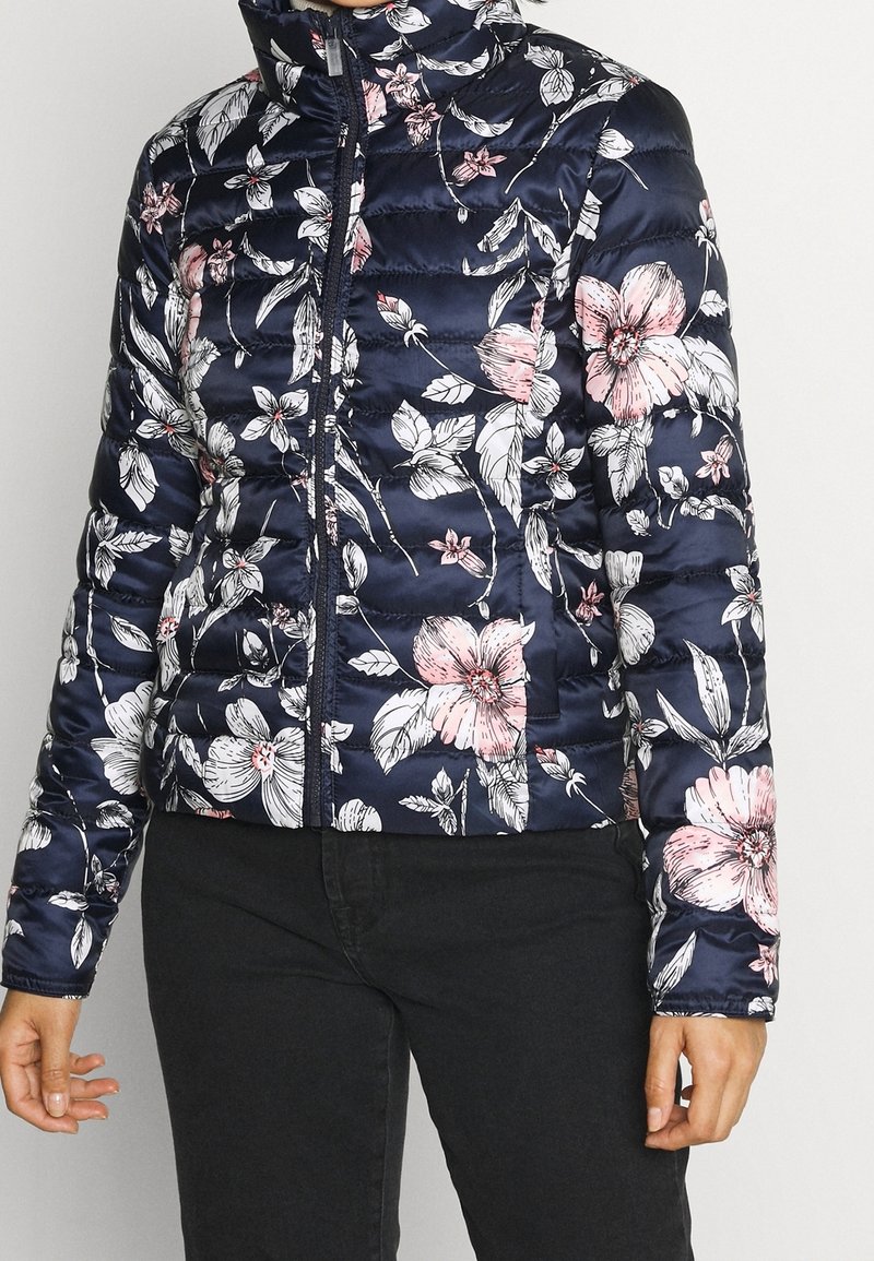Veste matelassée bleu marine avec imprimé floral rose et blanc, fermeture éclair devant, portée avec un pantalon noir.