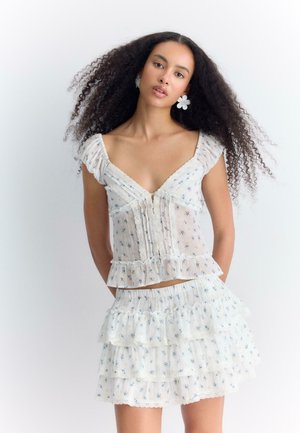Femme aux longs cheveux bouclés porte un chemisier blanc à volants fleuris et une jupe à volants avec des boucles d’oreilles en forme de fleurs, debout devant un fond uni.