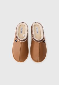 Brązowe skórzane kapcie z miękkim, kremowym włosiem w środku. Posiadają dekoracyjny, przeszyty brzeg oraz małą metkę z boku.