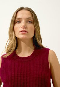 Ribbad stickad ärmlös tröja i djup vinröd, med rund halsringning och en texturerad yta med en åtsittande design.