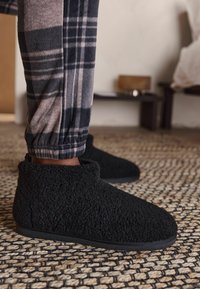 Papuci din lână texturată, negri, cu un design scăzut la gleznă. Suprafață moale și talpă solidă neagră, asortați cu pantaloni în carouri gri și negri.