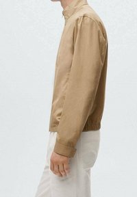 Beige skjorte med opretstående krave, elastisk talje, lange ærmer og glat tekstur, vist på en person mod en lys baggrund.