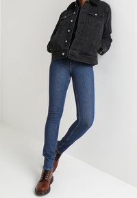 Zwarte denimjas met knoopsluiting, twee borstzakken, gecombineerd met blauwe skinny jeans en bruine veterschoenen. Eenvoudige, casual outfit.