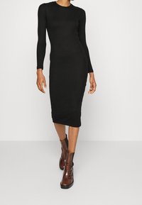 Schwarzes geripptes Strickkleid mit langen Ärmeln, taillierter Silhouette, knielangem Saum, kombiniert mit braunen Ankle Boots mit abgerundeter Zehenpartie und schwarzer Sohle.