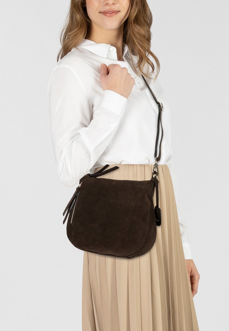 Borsa a tracolla in suede marrone con una forma curvata, tasche con zip e una finitura texturizzata, indossata su una gonna plissettata beige.