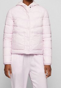 Giacca imbottita rosa chiaro con design corto, cuciture orizzontali, bottoni a pressione e due tasche frontali. Dotata di cappuccio con fodera bianca.