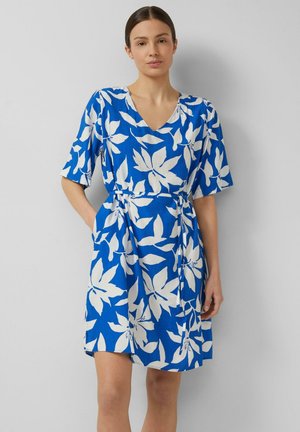 Kobieta w niebieskiej sukience za kolano z dużym białym kwiatowym wzorem, dekoltem w serek, krótkimi rękawami i wiązanym paskiem w talii, stojąca na szarym tle.