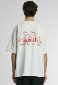 Tricou alb din bumbac cu un design grafic roșu pe spate, reprezentând o clădire și textul "Ciao Amore" și "Interzis, Casa Moda Milano."