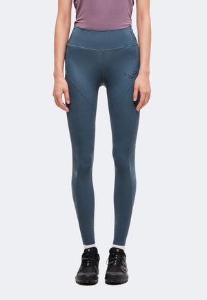 Kvinde iført blå højtaljede træningsleggings med et logo på låret og sorte trail løbesko med hvide sokker.