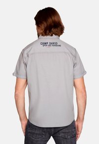 Camisa de manga corta de color gris claro con dobladillo recto y texto bordado en la parte posterior que dice "CAMP DAVID OPEN SEA KAYAKING". Textura suave.