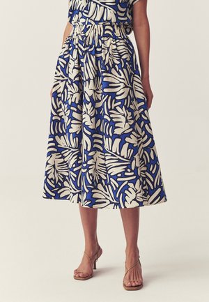 Kobieta w niebiesko-białej spódnicy midi z tropikalnym liściastym wzorem, z elastycznym paskiem w talii, nosząca beżowe sandały na wysokim obcasie z paskami.