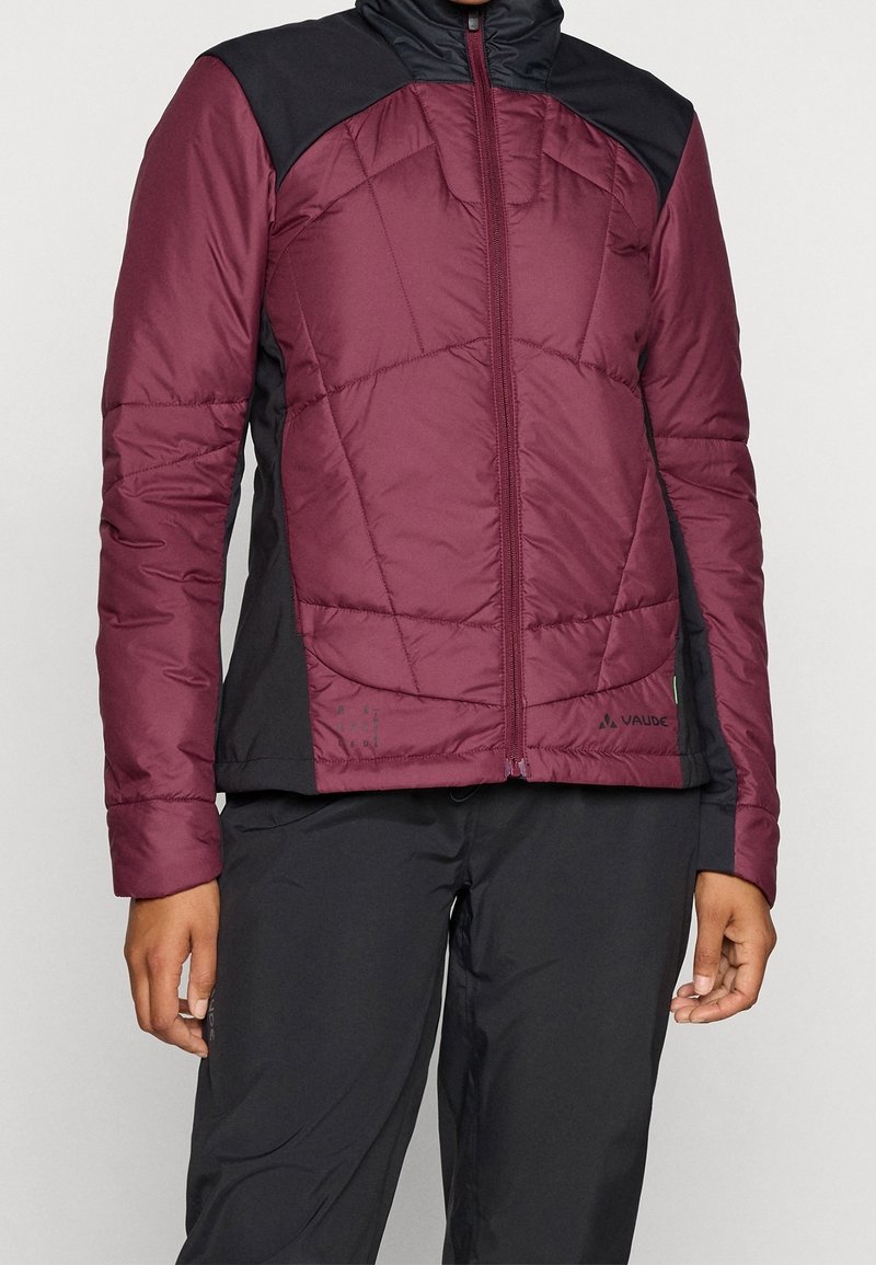 Femme portant une veste matelassée bordeaux et noire avec une fermeture éclair à l'avant et un pantalon noir, debout devant un fond uni.