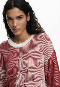 Roter und cremefarbener gestrickter Pullover mit Wellenmustern, geripptem Kragen und weiten Ärmeln. Die strukturierte Stoffqualität verleiht visuelles Interesse.