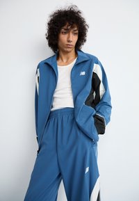 Person med krøllet hår iført en hvit tanktop under en blå og svart zip-up jakke og matchende blå treningbukser, stående mot en hvit vegg.