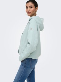 Femme portant une veste à capuche bleu clair et un jean bleu, debout de profil, les mains dans les poches de la veste, sur un fond uni.
