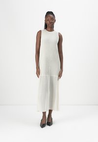Sleeveless cream maxi dress with ribbed texture, flowing bottom, and a slight sheen. Worn with black pointed-toe heels.