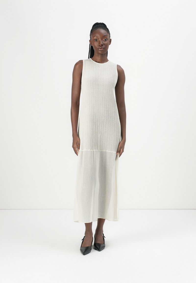 Sleeveless cream maxi dress with ribbed texture, flowing bottom, and a slight sheen. Worn with black pointed-toe heels.