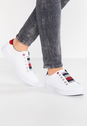 Persona que lleva vaqueros grises desgastados y zapatillas blancas sin cordones con rayas de purpurina en rojo, plata y azul marino sobre un fondo blanco.