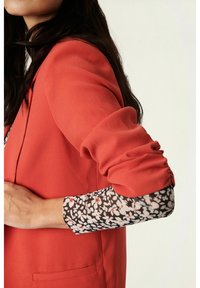 Blazer corallo con design aderente, caratterizzato da maniche arricciate e un polsino a fantasia floreale contrastante in nero, bianco e rosso.