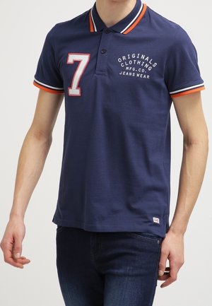 Mand iført marineblå polo med orange og hvide striber på krave og ærmer, nummer 7 og teksten "ORIGINALS CLOTHING" på brystet.