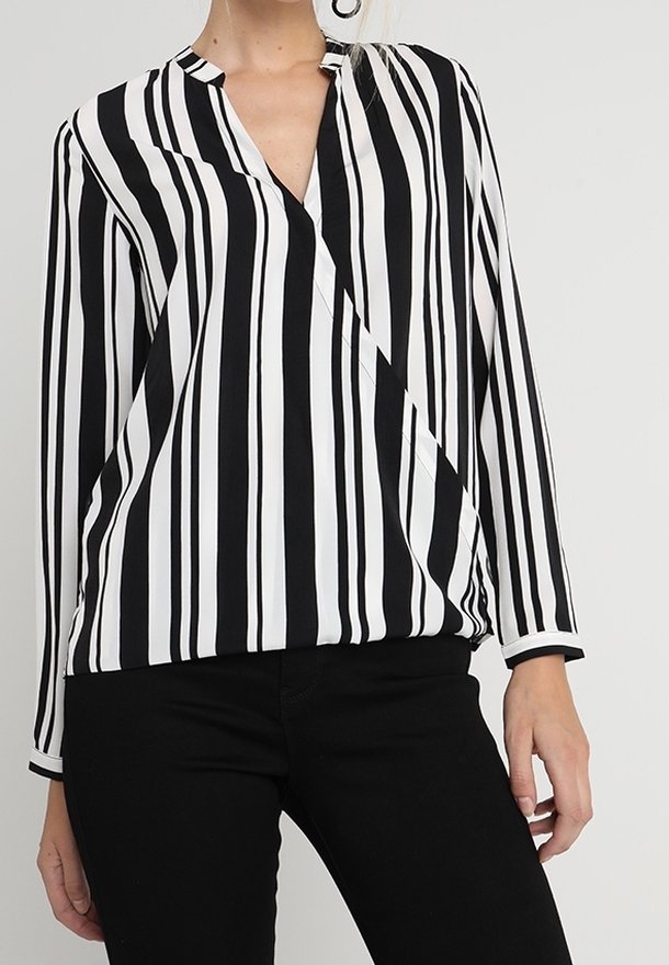 Femme portant un chemisier à rayures verticales noires et blanches à manches longues avec un col en V et un pantalon noir sur un fond uni.
